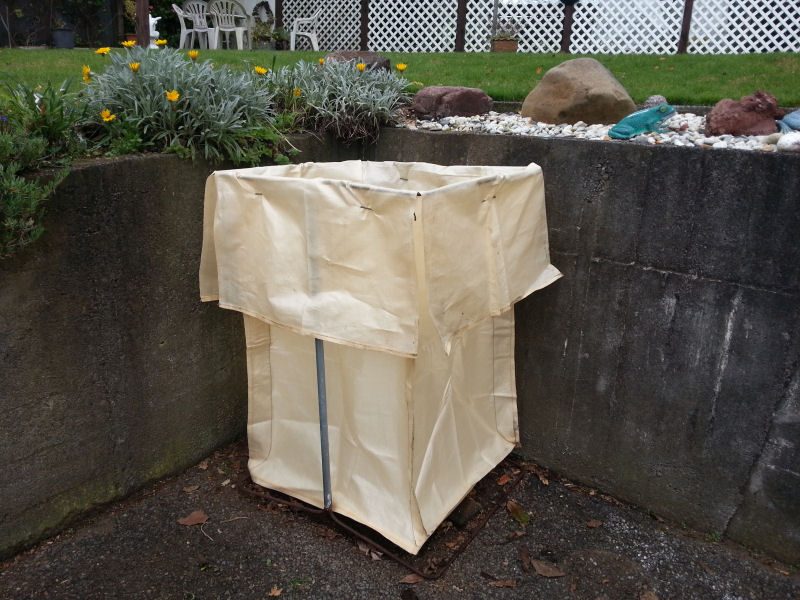 A Greenawaste 450L garden bag held open in its steel frame, set up beside a garden wall — the bag sits wherever suits you, not at the kerb
